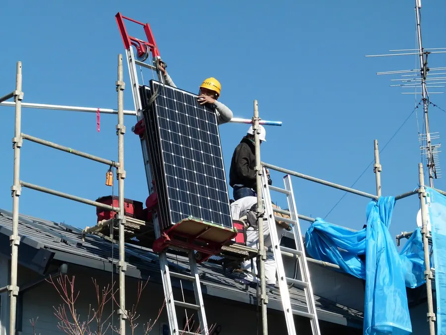 Dos instaladores con casco subiendo un panel solar por un andamio hasta el techo