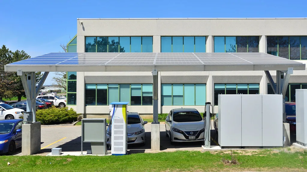 Electrolinera con techo solar, un cargador rápido y uno de pared frente a un edificio de oficinas